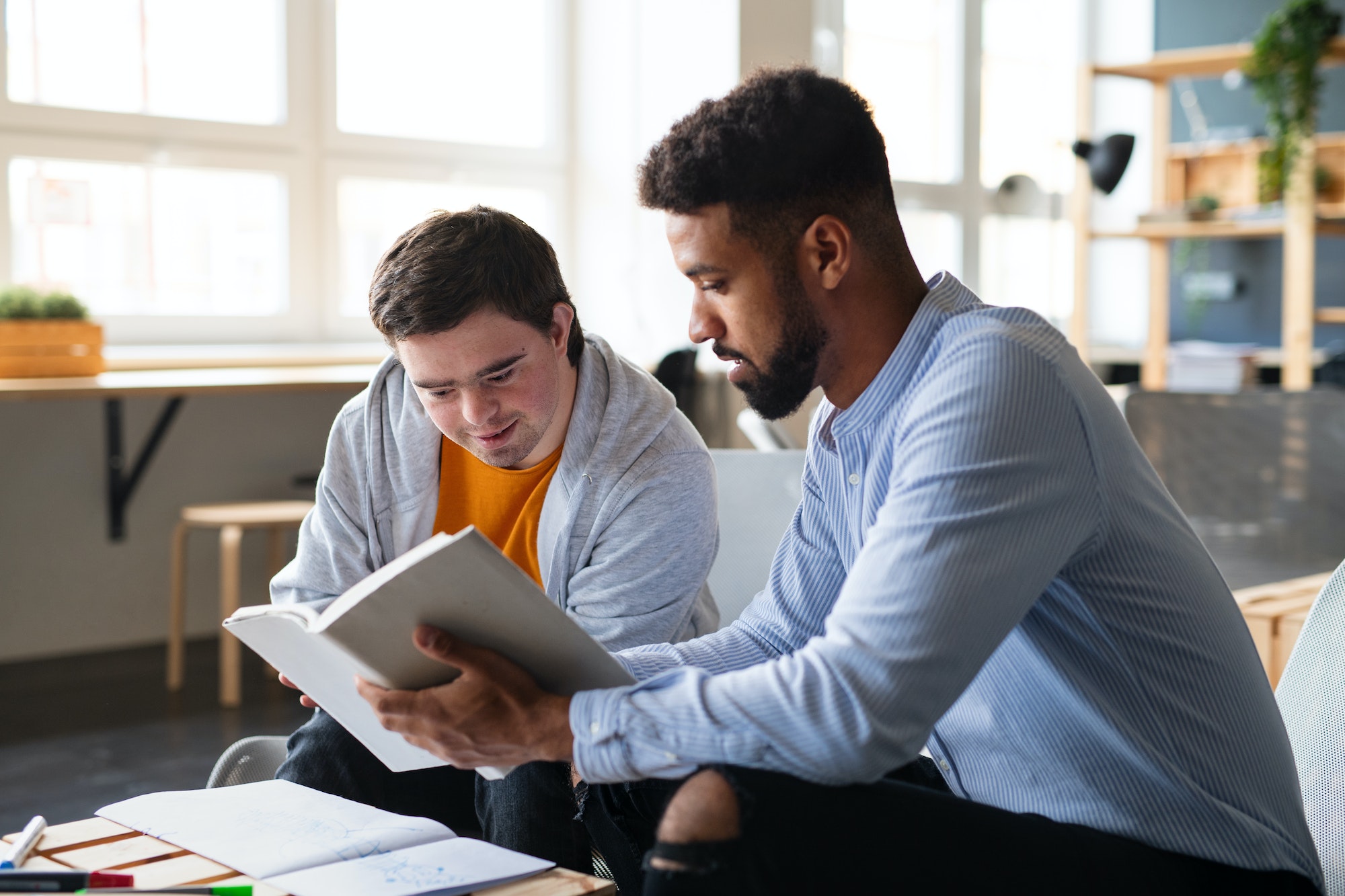 Young happy man with Down syndrome and his tutor studying indoors at school